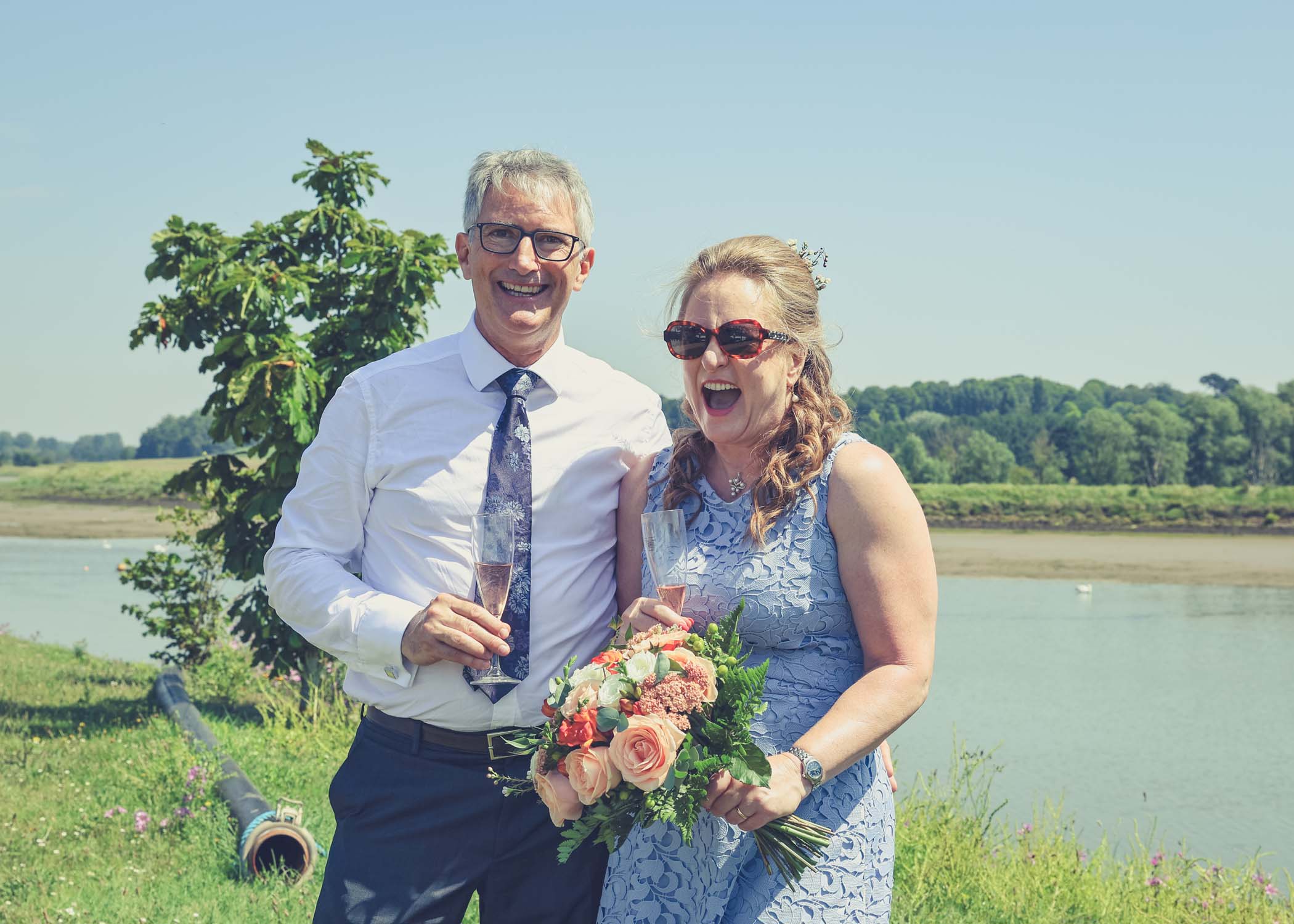 A bride in sunglasses and the groom laughing on the penisula at woodbridge marina on a wedding day in Woodbridge captured by Suffolk Wedding Photographer Hayley Denston Photography